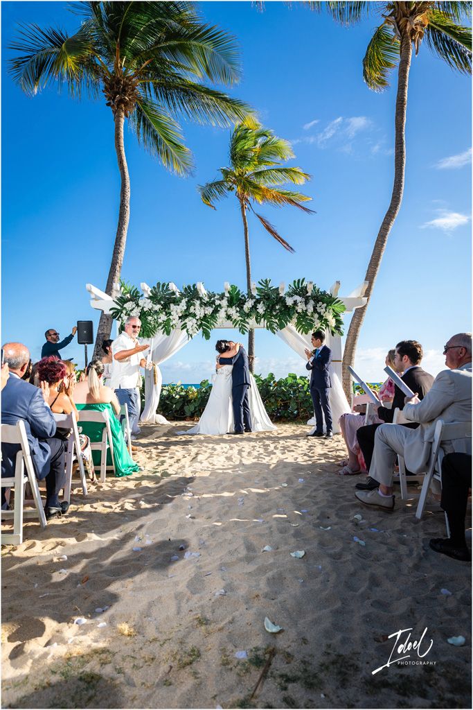 A beach wedding ceremony setup with palm trees and floral arch while guests watch from white chairs on the sand.