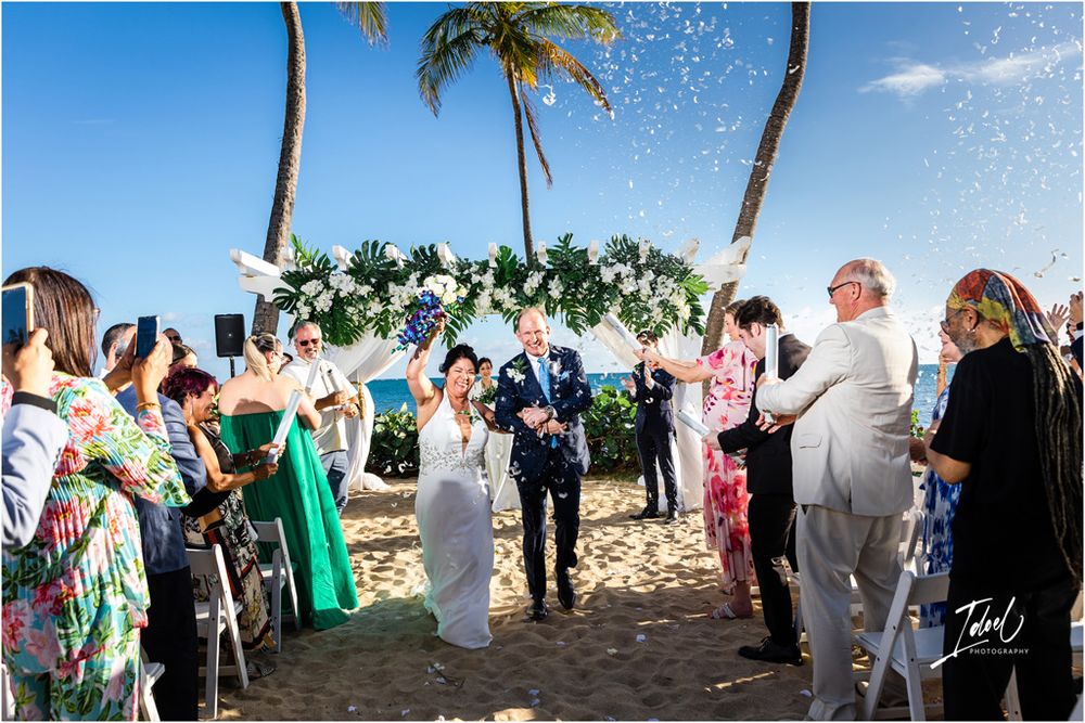 Wedding guests celebrate as bubbles float through the air during an outdoor beach ceremony.