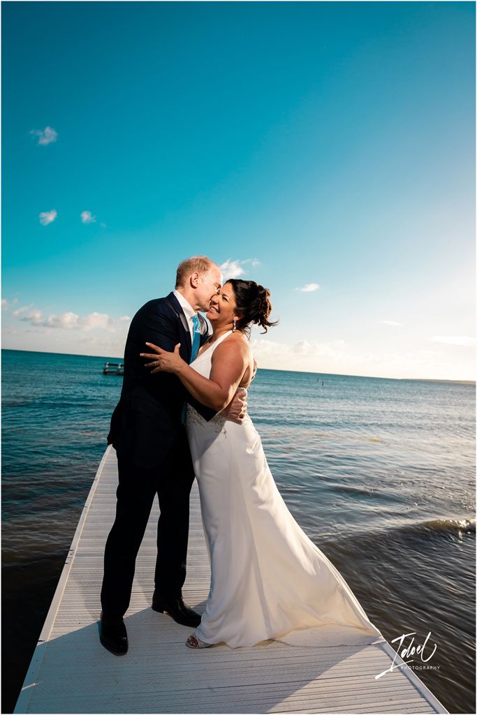 A joyful wedding moment captured on a white dock extending into bright blue ocean waters.