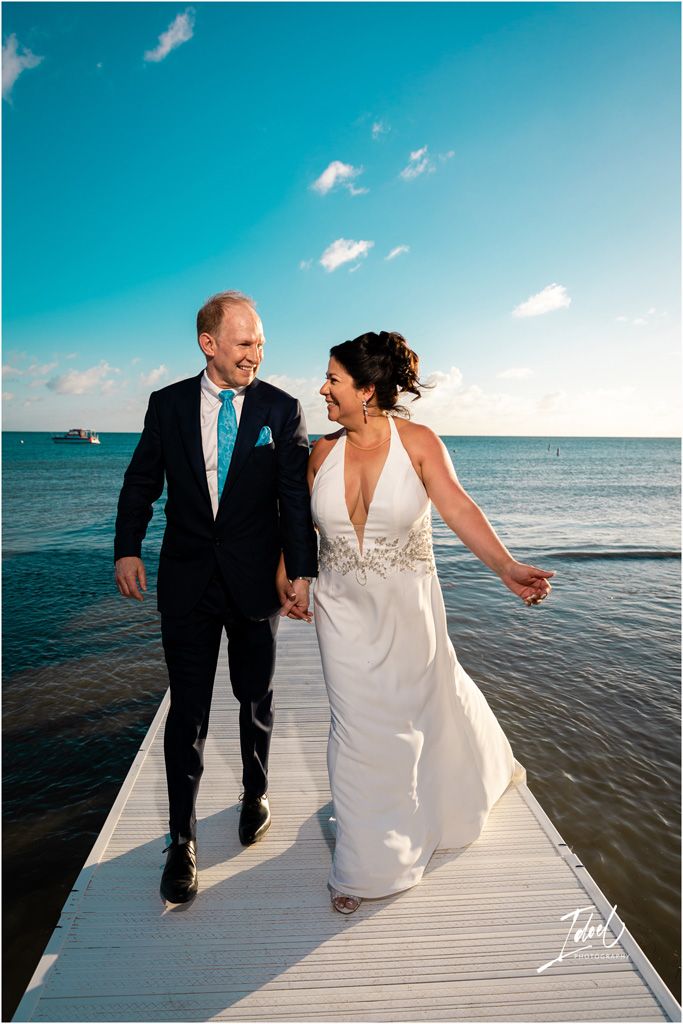 A couple walking hand in hand on a white boardwalk with the ocean and bright blue sky behind them.