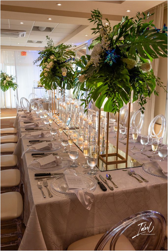 An elegant wedding reception table with tall tropical greenery centerpieces and formal place settings.