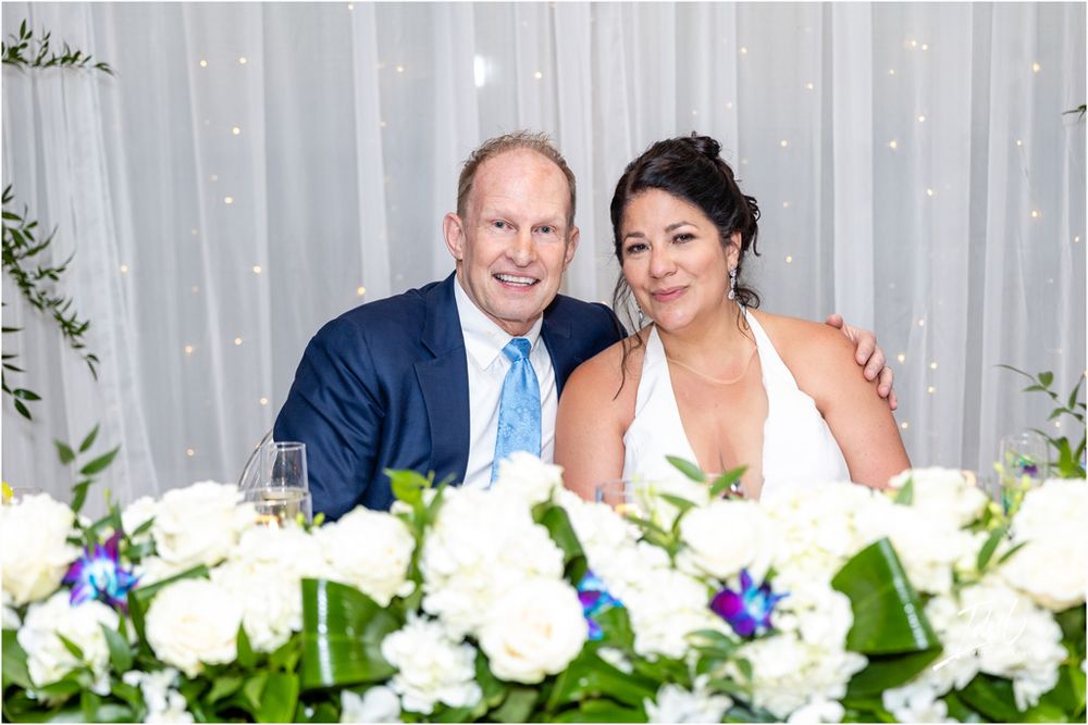 Newlyweds seated at wedding reception table decorated with white floral arrangements.
