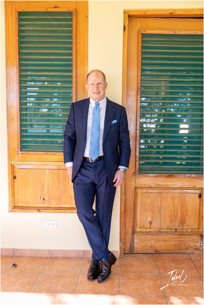 A person in a navy suit and blue tie poses against wooden doors with green shutters.