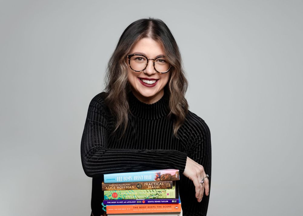 Woman in black turtleneck sweater holds stack of colorful books while smiling.
