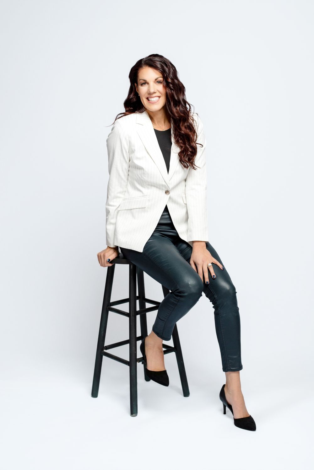 Professional portrait showing person in white blazer and dark pants sitting casually on black bar stool against light background.