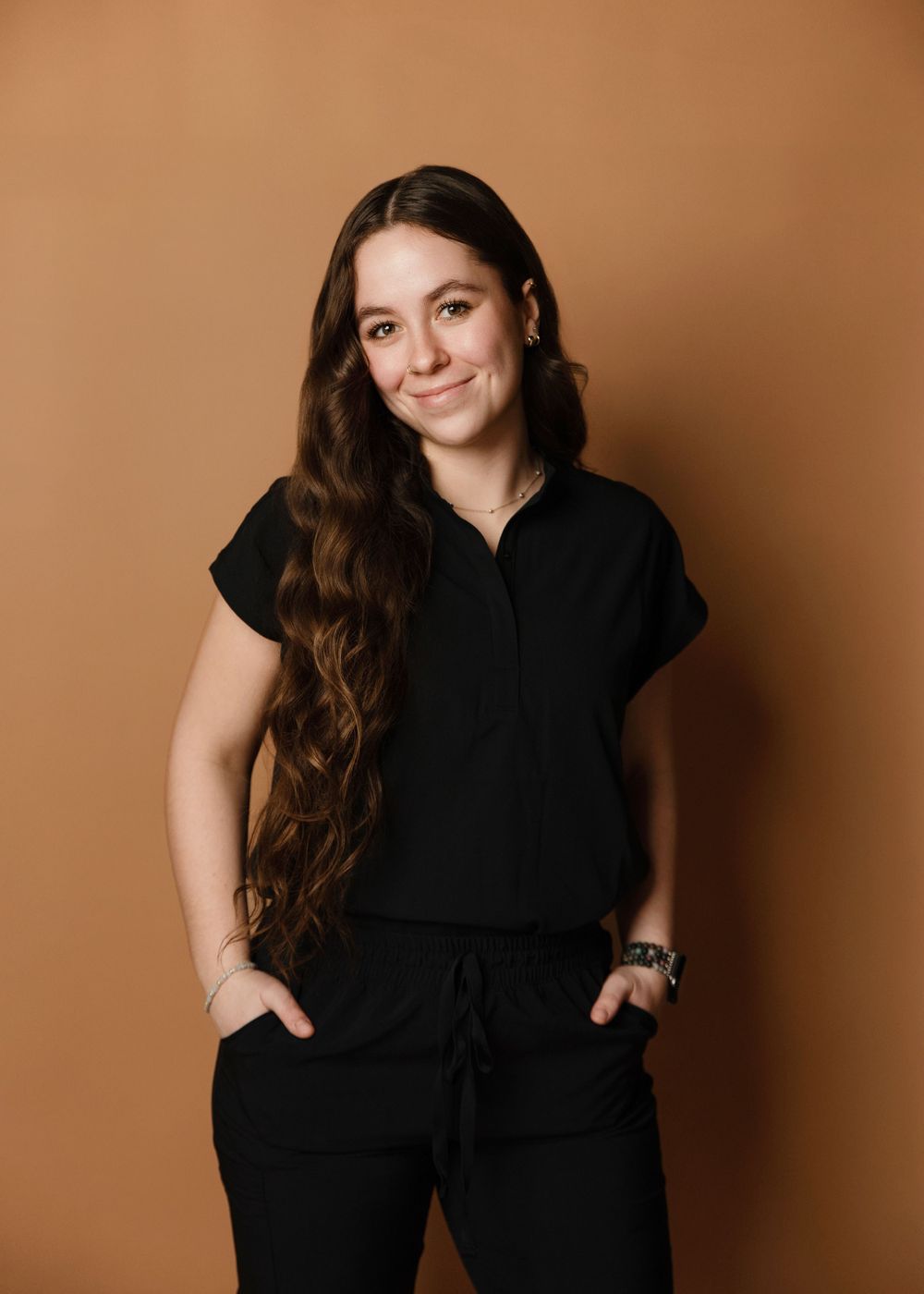 Portrait of someone wearing an all-black casual outfit with long brown hair against a brown backdrop.