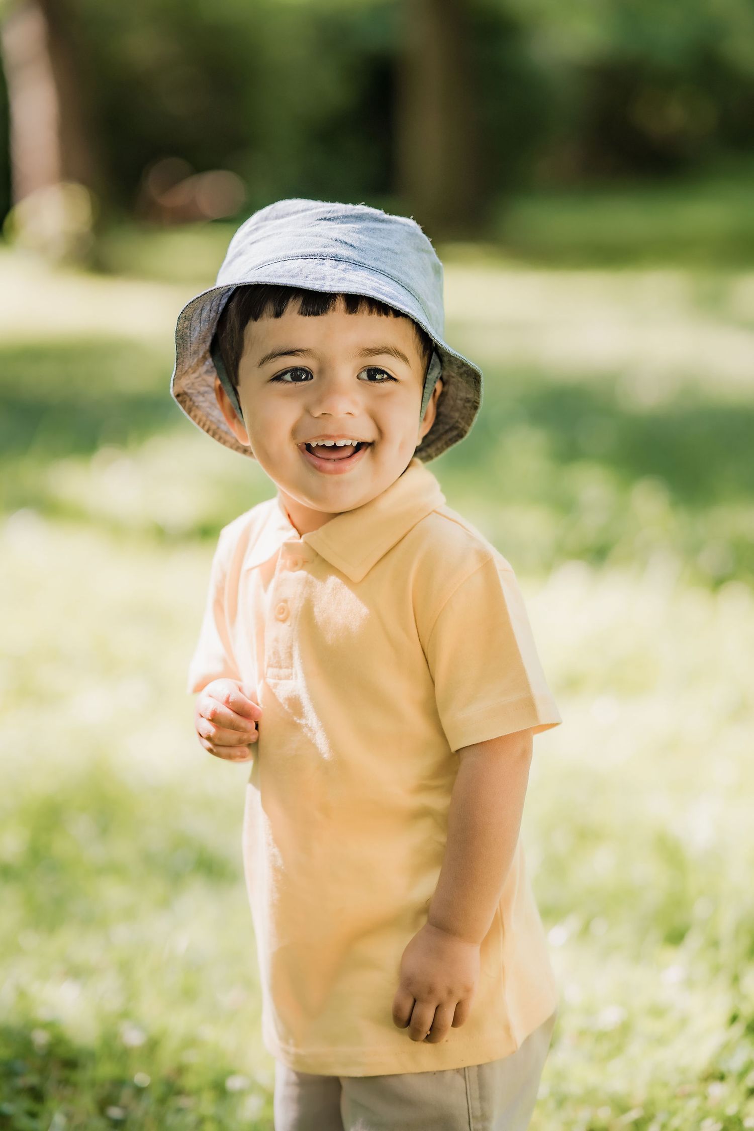 Joyful toddler wearing a blue bucket hat and peach t-shirt smiles outdoors on a sunny day.