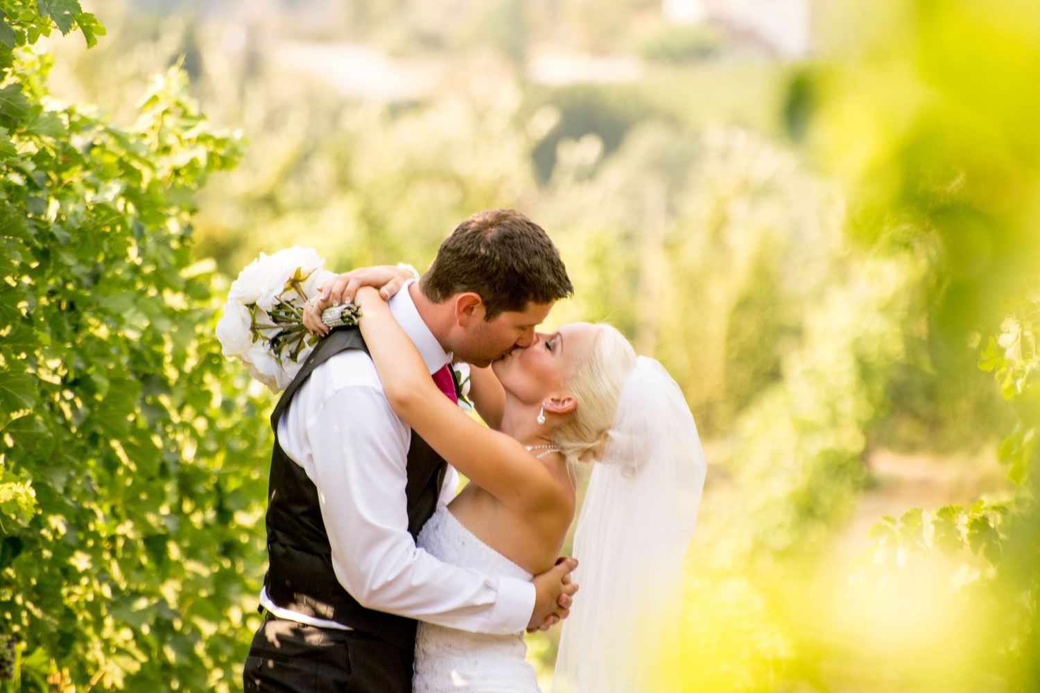 Romantic wedding couple embracing in sunlit vineyard with soft focus greenery.