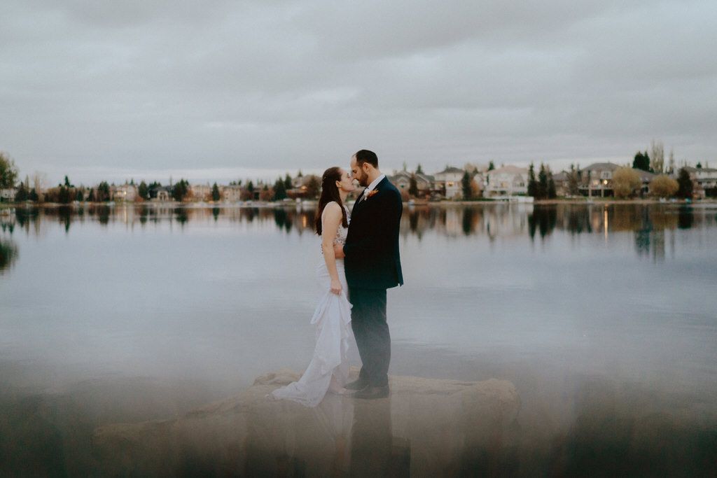 Couple standing together by a reflective lake with buildings in the background.