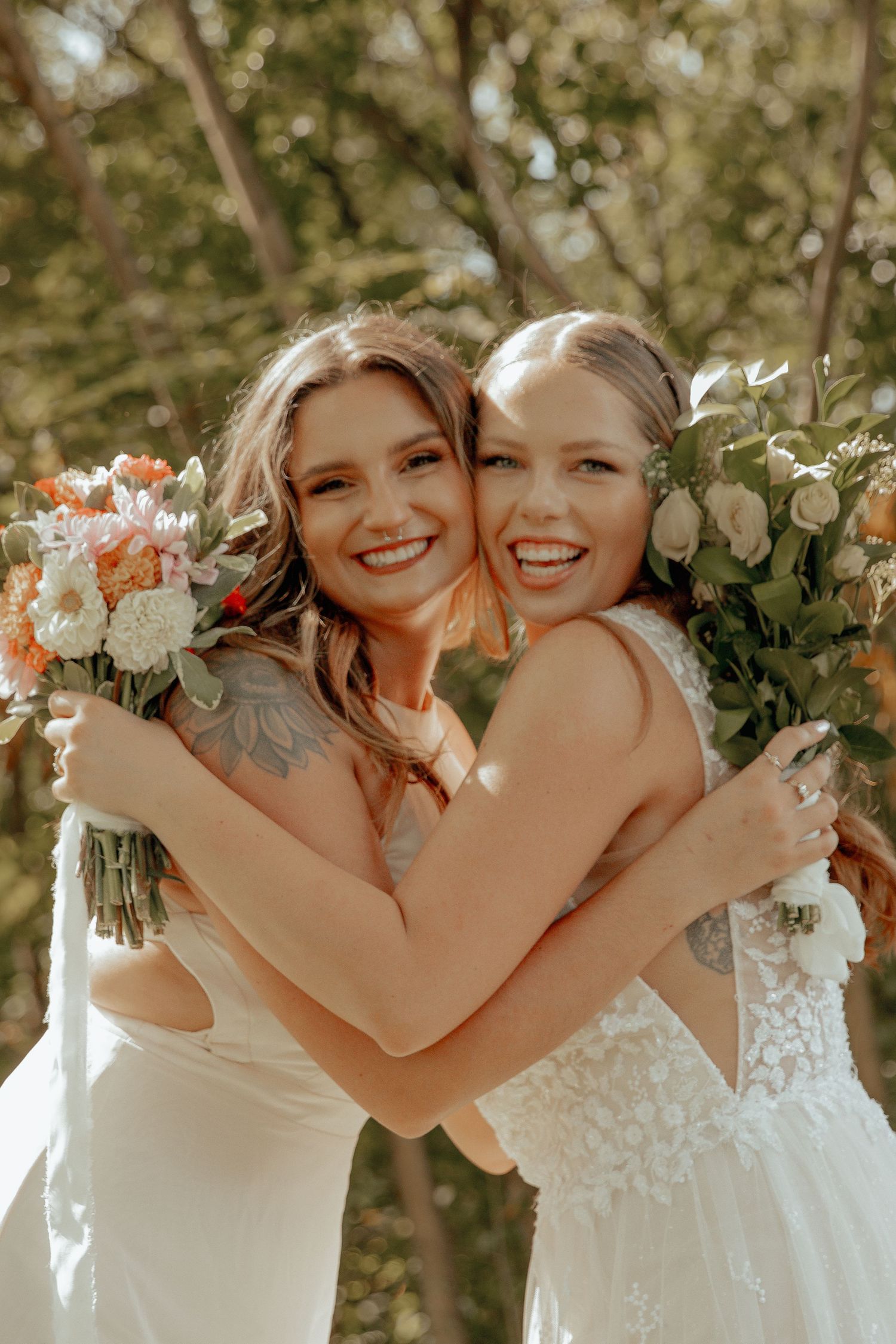 Two people in white wedding dresses embrace while holding bouquets in outdoor setting.