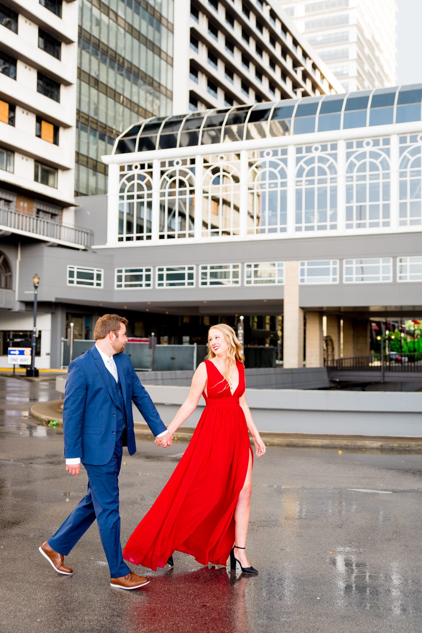 blonde woman in red dress holds hand leading fiance in navy suit outdoors