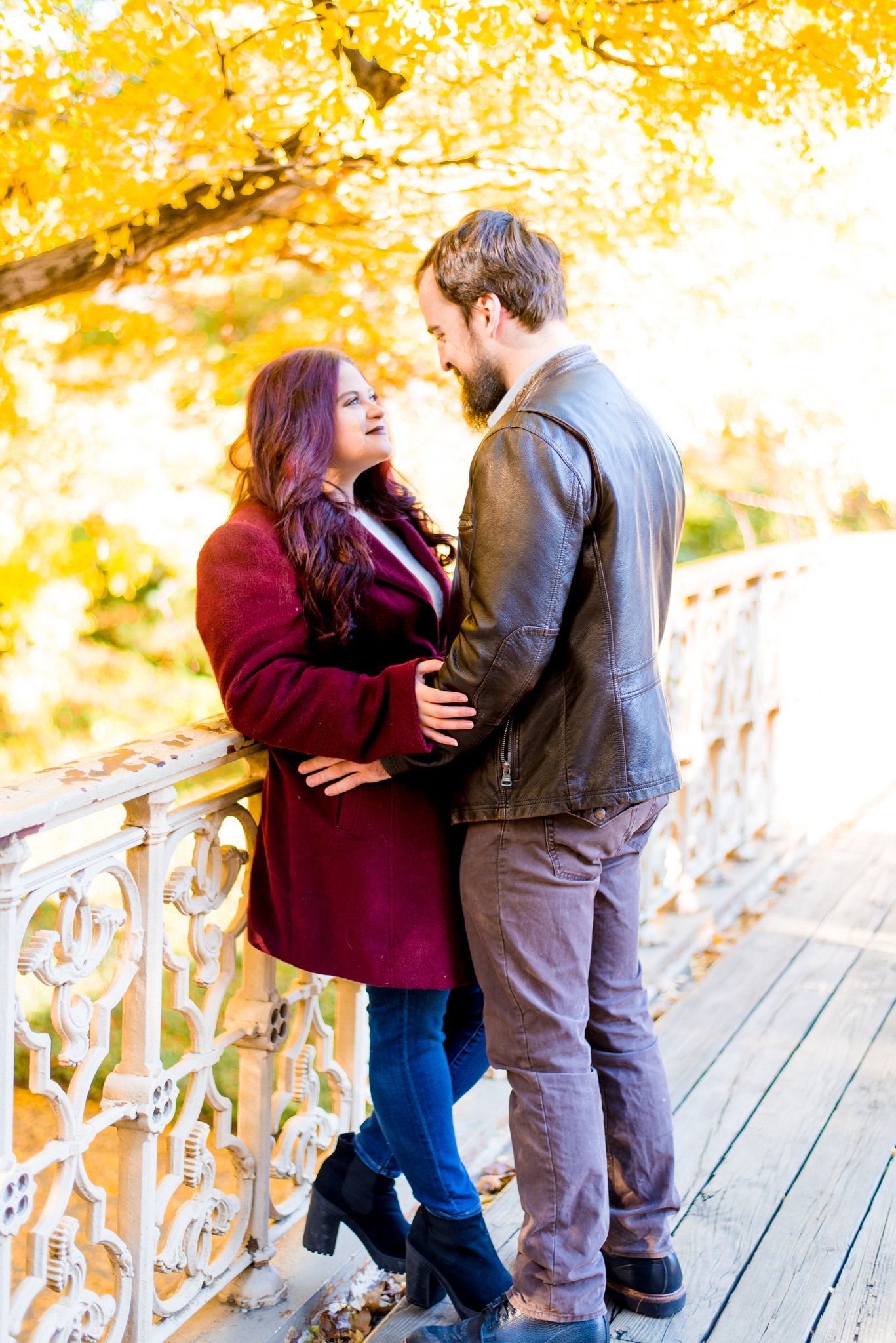 woman in cranberry jacket leaning against Pinebank Arch in Central Park in November with fiance in brown leather coat