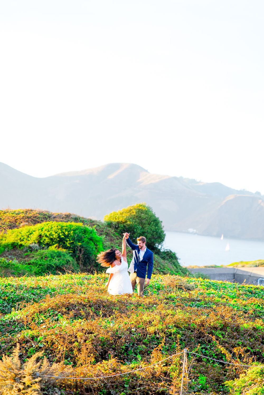 man in navy suit twirling woman in white dress at sunset in hills in front of Golden Gate Bridge in background