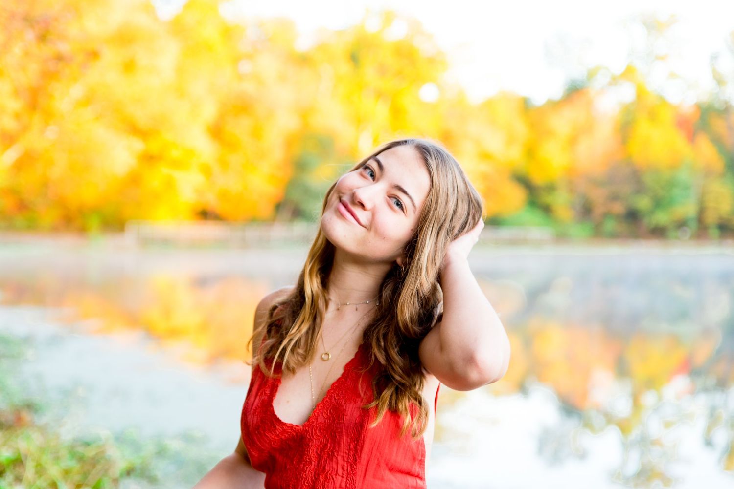 young woman in orange shirt plays with hair and smiles in front of a pond in Chicago with yellow trees behind her