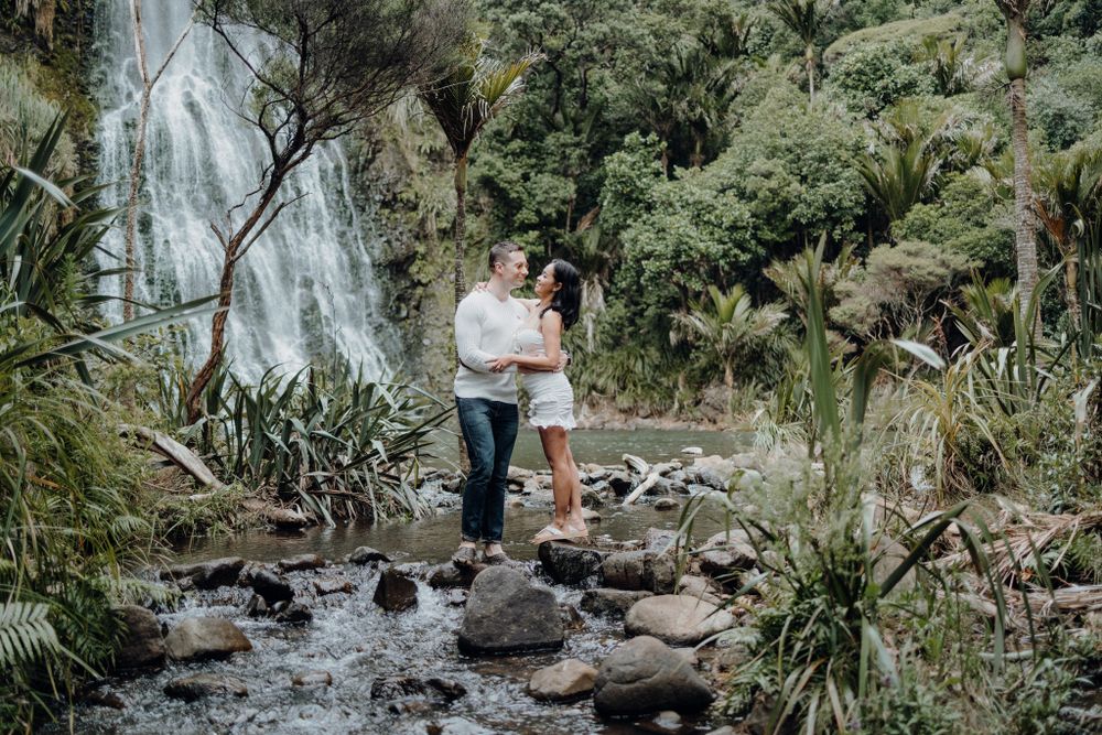A couple stands together on rocky stream bed with waterfall and tropical foliage.