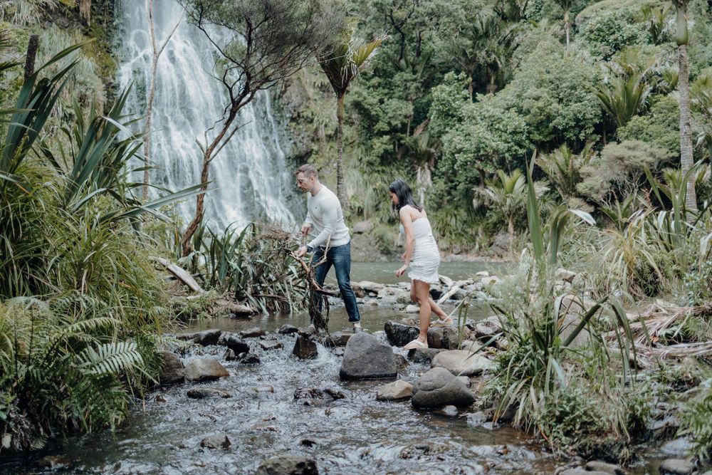 A couple traverses a rocky stream bed surrounded by lush tropical vegetation.