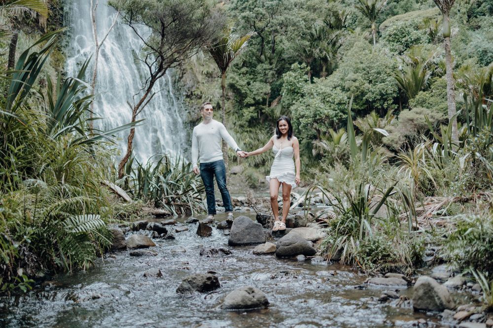 A couple explores a rocky stream bed with a magnificent waterfall in the background.