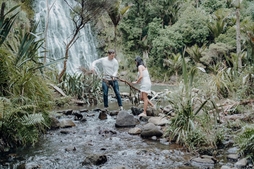 Two people navigate across rocky terrain near a flowing stream with a waterfall view.