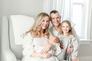 Family of four all smile as mother holds newborn baby and father hugs toddler girl as they sit in the nursery