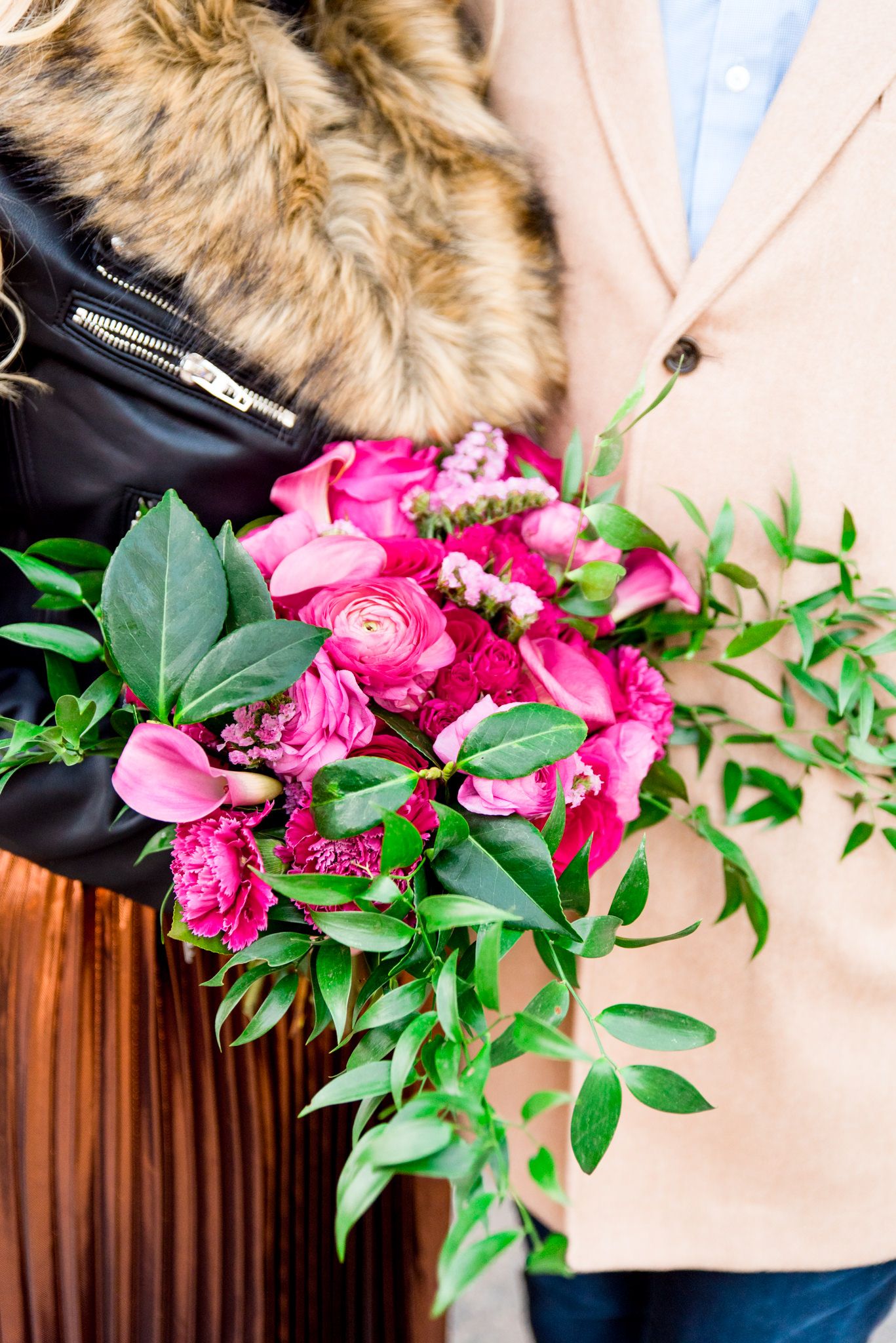 pink roses and ranunculus bouquet held by woman in black and copper outfit and man in tan coat