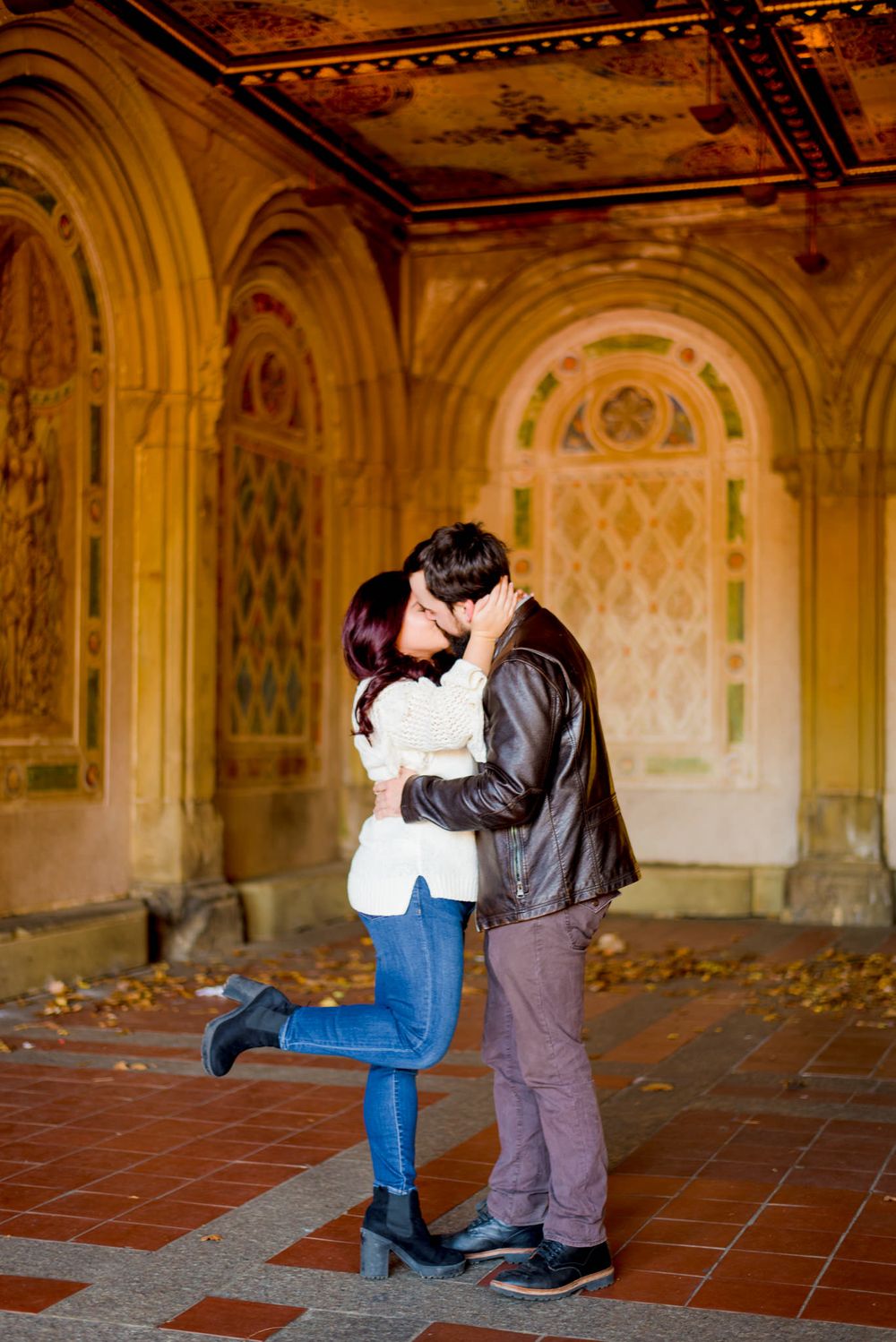 man in brown jacket and woman in white sweater kissing and hugging at Bethesda Terrace in Central Park for engagement