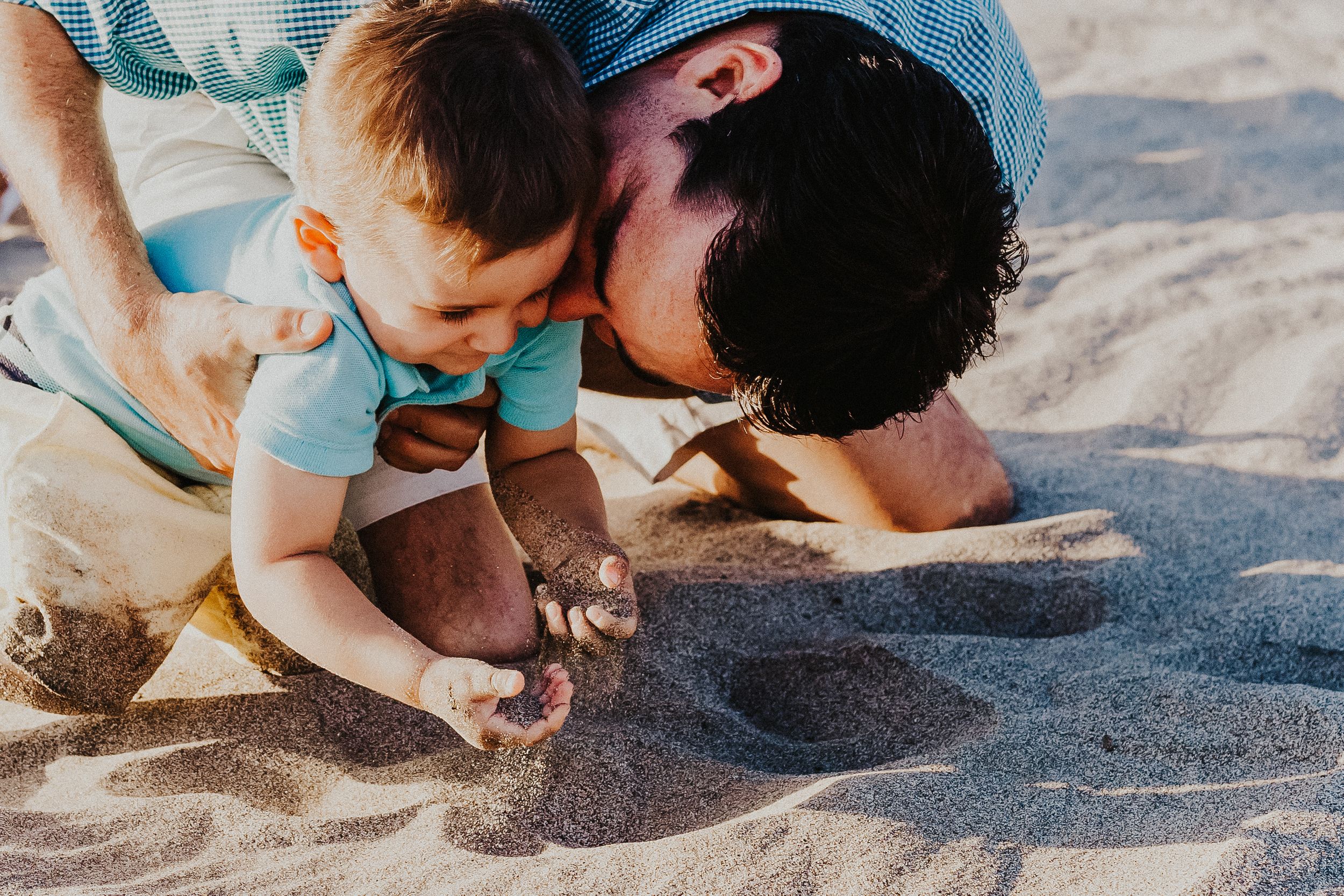 dad kisses son's cheek as he plays in the sand on the beach