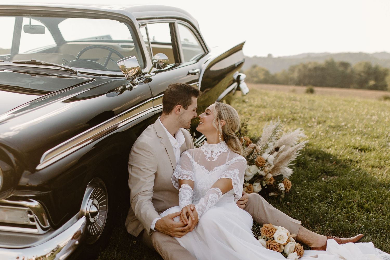 Newlyweds share an intimate moment while sitting against their classic black car in a field.