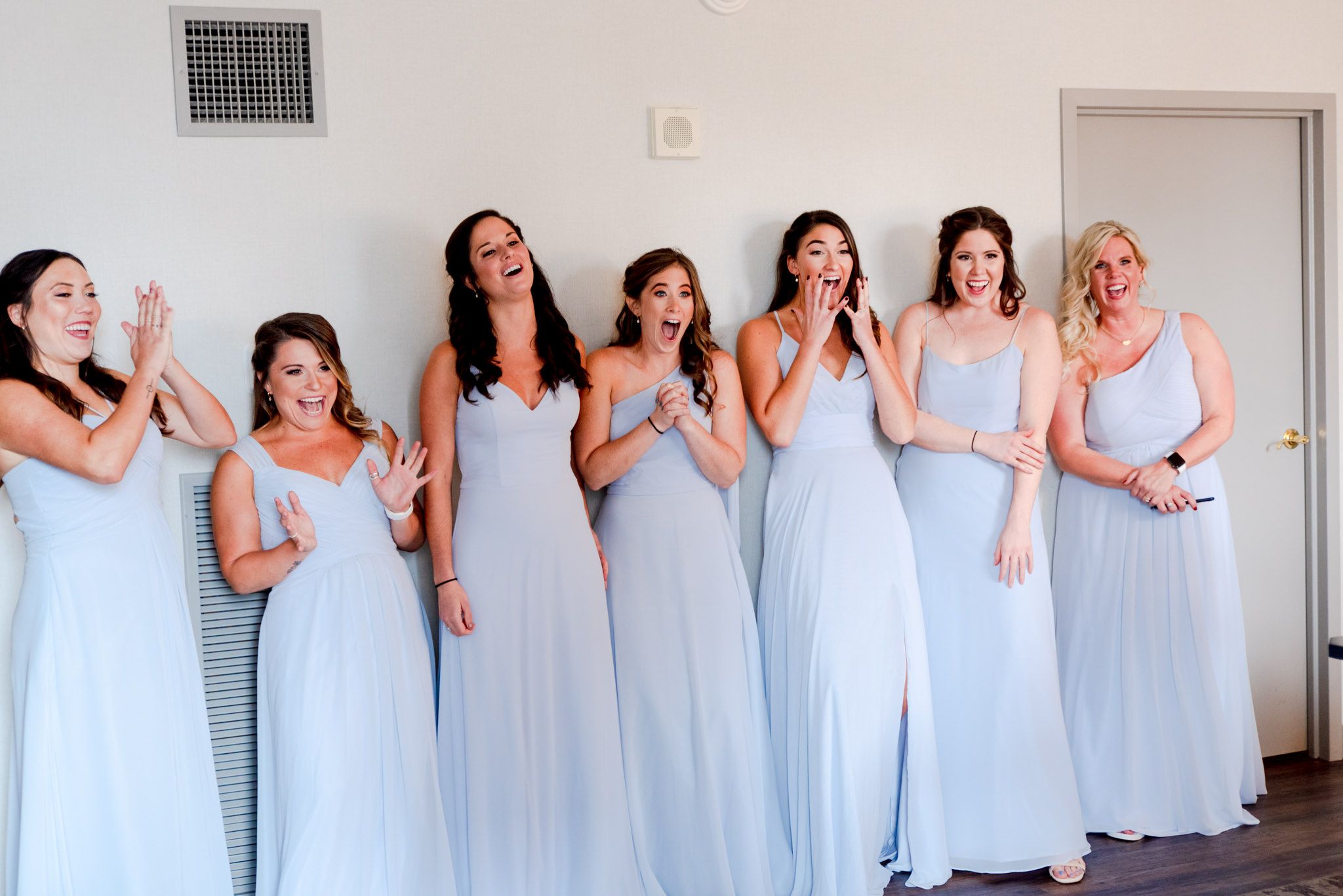 7 bridesmaids in light blue dresses smiling and clapping after seeing the bride for the first time on wedding day