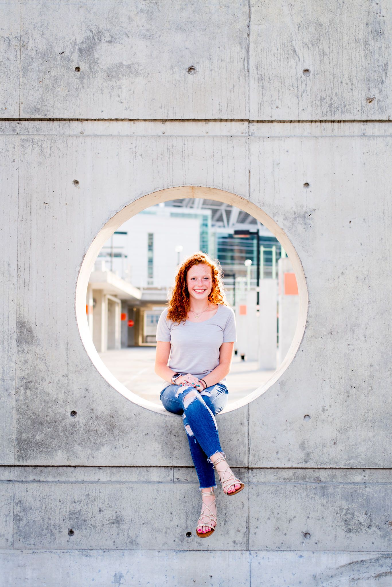 young woman with curly red hair sitting in a circle cut out from concrete wall in parking structure, smiling