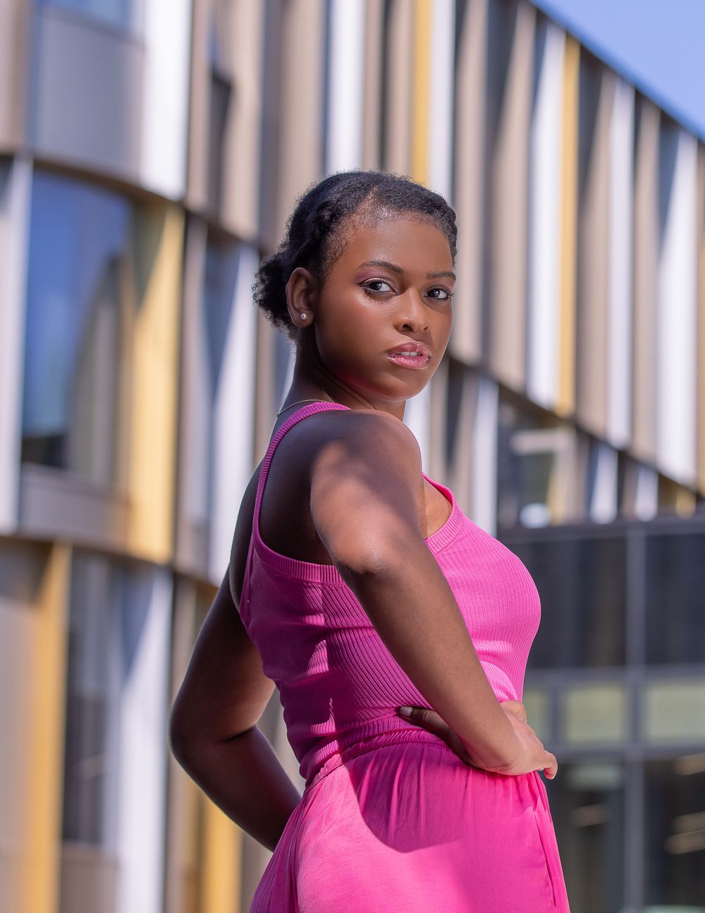 A black woman wearing a bright pink dress looks over her shoulder outside a college building