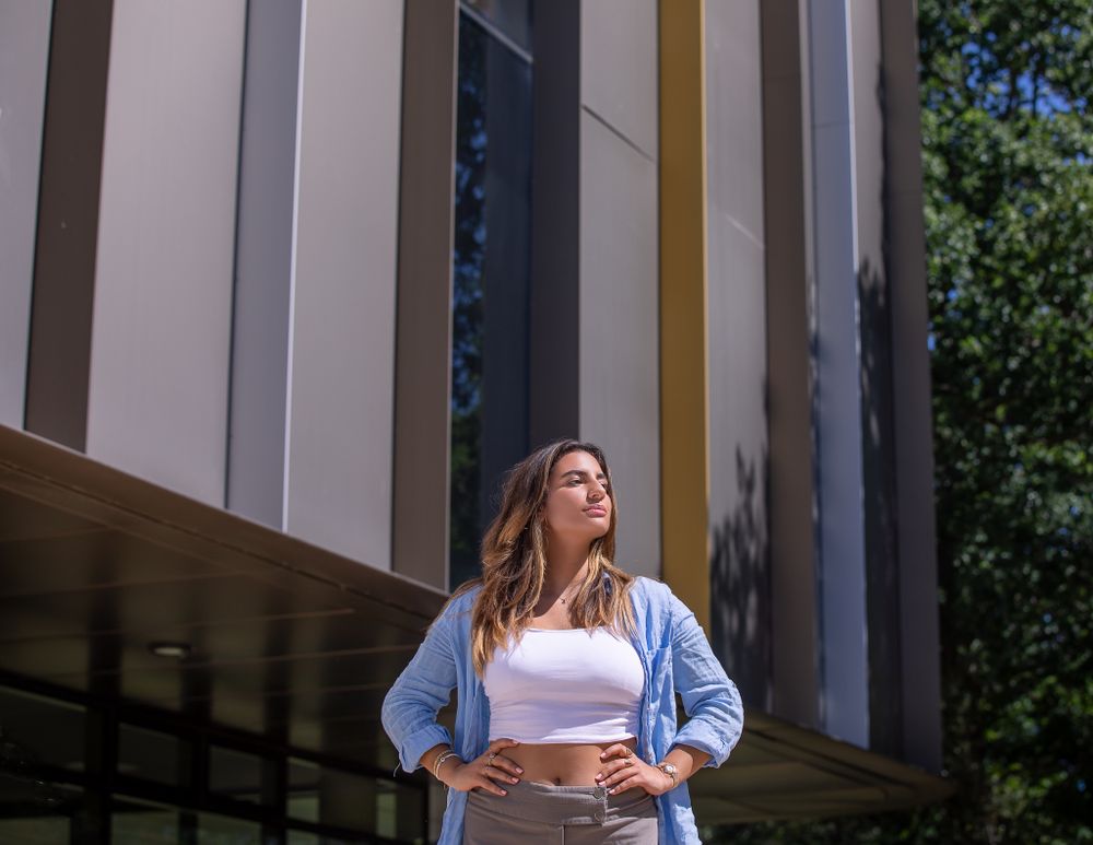 A woman wearing a white t shirt and denim short stands with her hands on her hips outside a large, modern building