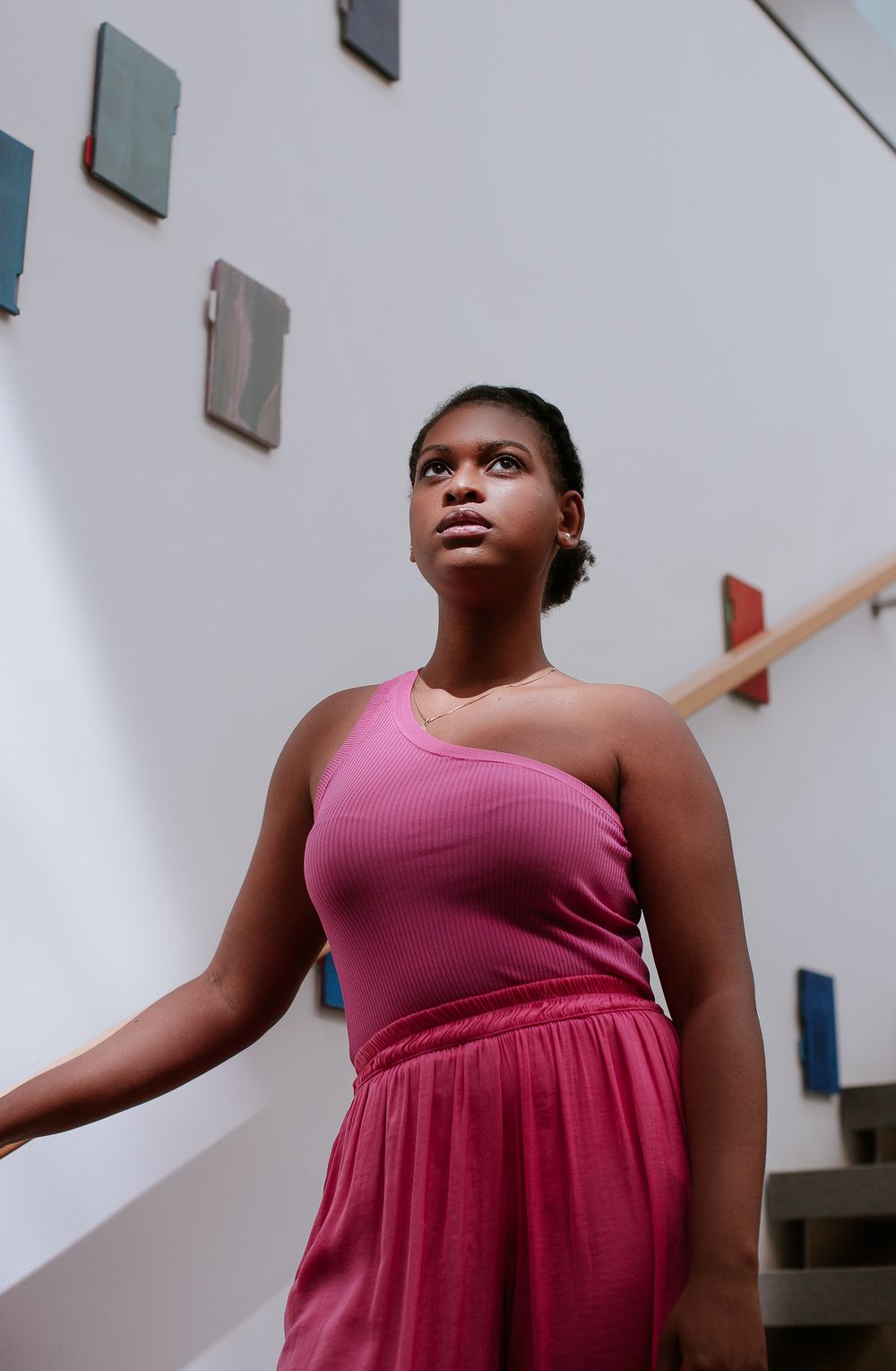 A black woman wearing a hot pink dress pauses and looks up whilst walking down a large staircase
