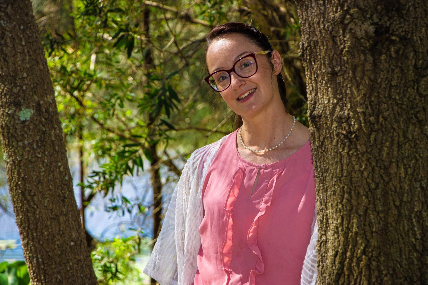 Outdoor portrait of someone in a pink shirt and grey cardigan standing between two trees.