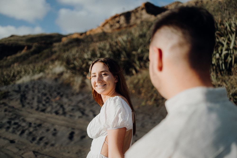 A couple poses against a rocky coastal backdrop in white outfits.
