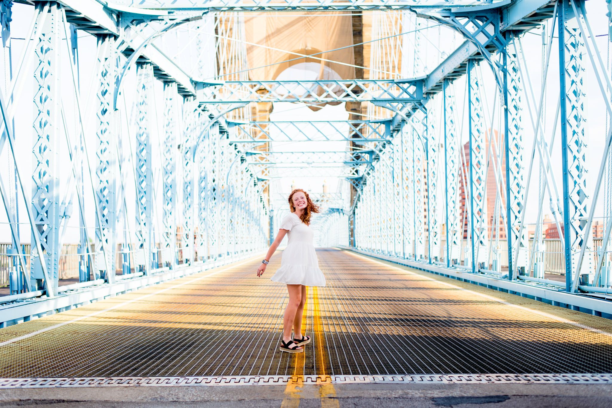 young woman in short white dress spinning around on blue Roebling Bridge for downtown Cincinnati senior pictures