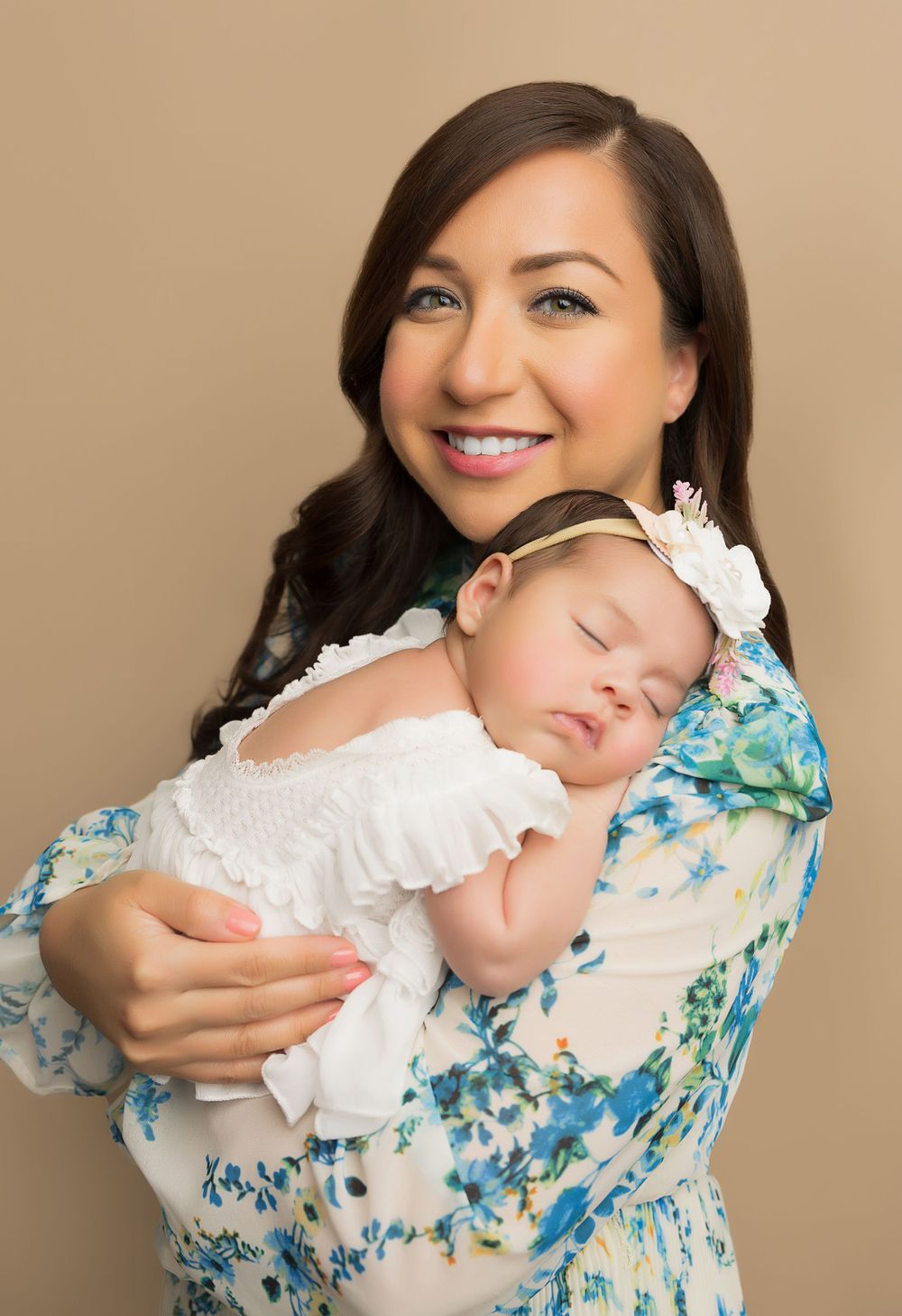 Woman in floral dress holding sleeping baby in white dress.