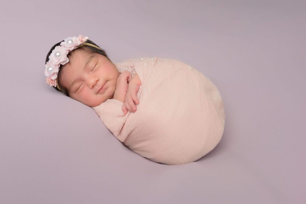 Peaceful sleeping newborn in beige wrap with floral headband against mauve backdrop.