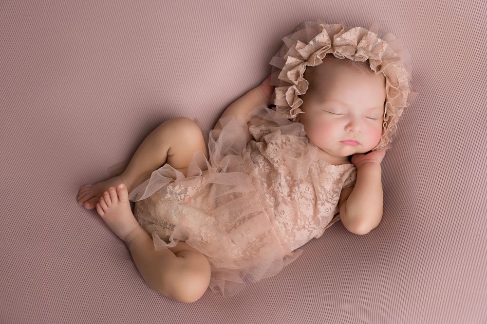 Sleeping newborn in delicate lace wrap and bonnet on pink background.