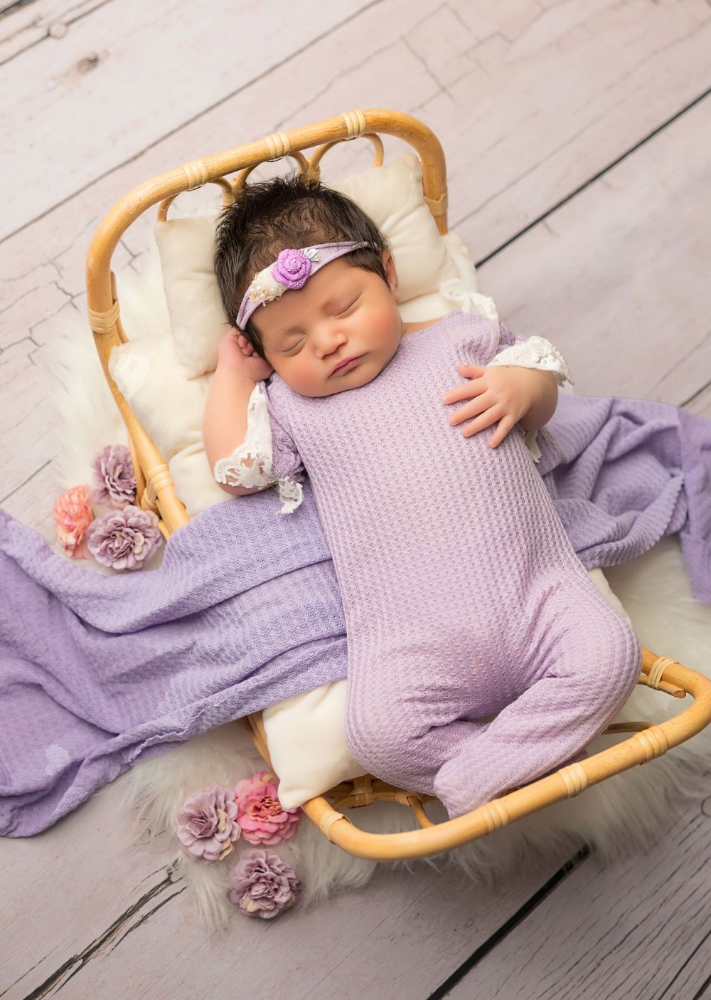 Sleeping baby in purple knit outfit resting on wicker bassinet with floral decorations.