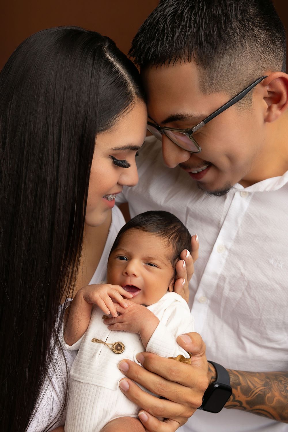 Intimate family portrait with newborn baby in white outfit against brown background.