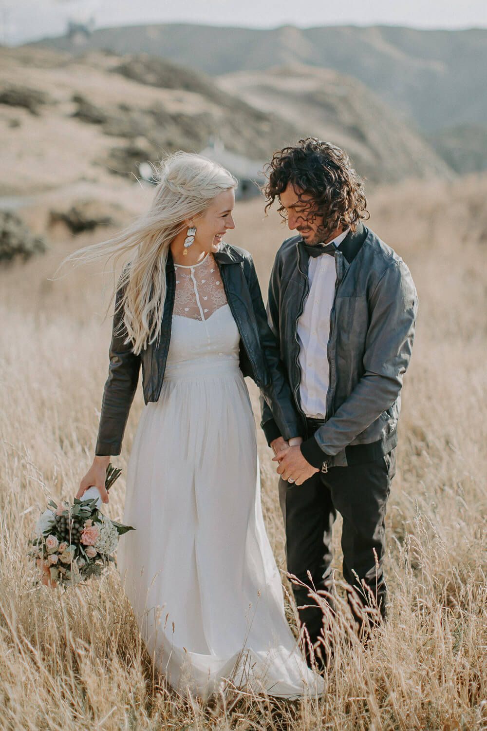 A couple in wedding attire stand together in a golden field with mountains in the background.