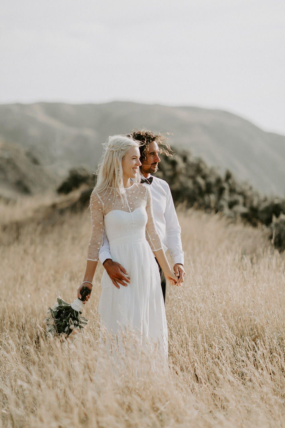 Wedding couple in white attire standing in golden grass with mountain backdrop.