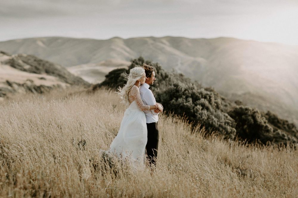Wedding couple embracing on hilltop with golden grass and mountain views.