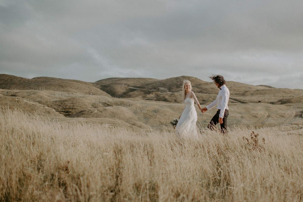 Wedding couple walking through tall grass with rolling hills in background.