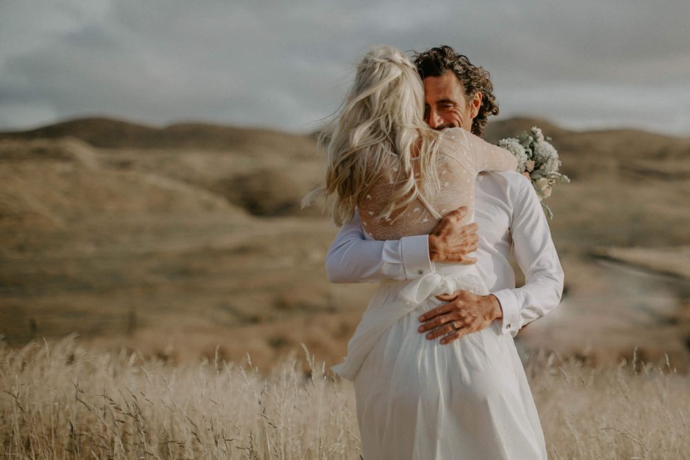 Intimate wedding embrace in golden grass field with hills in background.