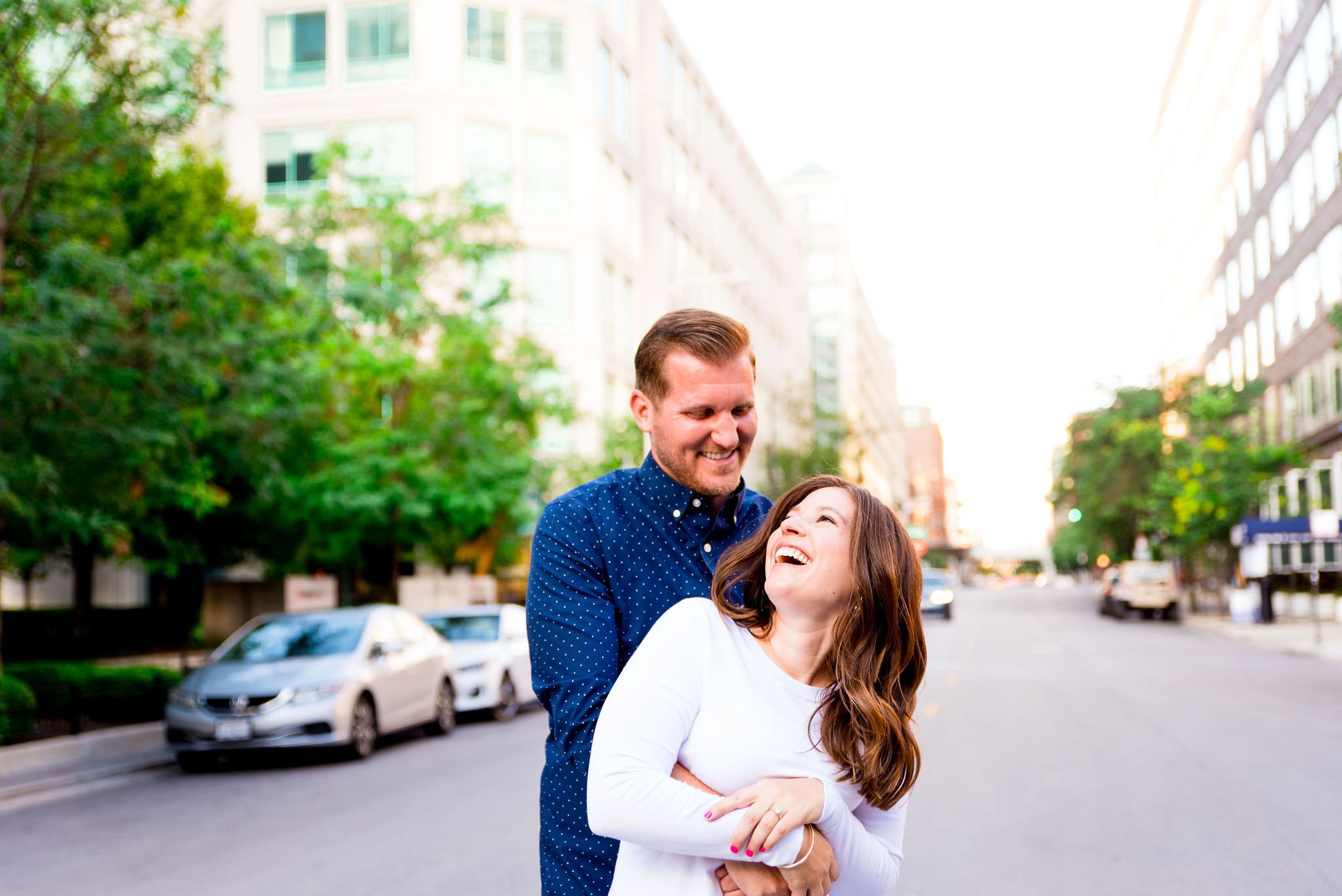 redhead man in navy shirt hugging woman in white sweater, laughing in the middle of street for sunset engagement picture
