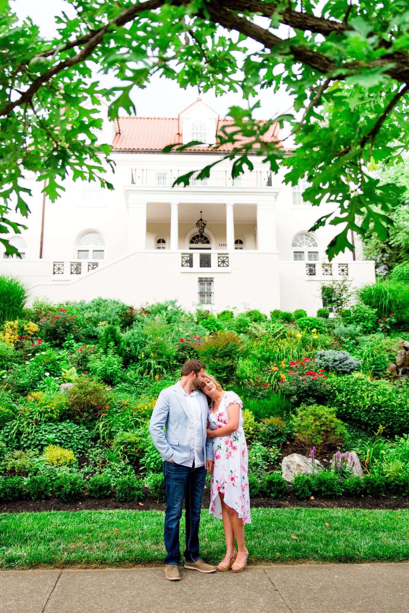 engaged couples in floral dress and light blue suit jacket and jeans standing in front of white Louisville mansion