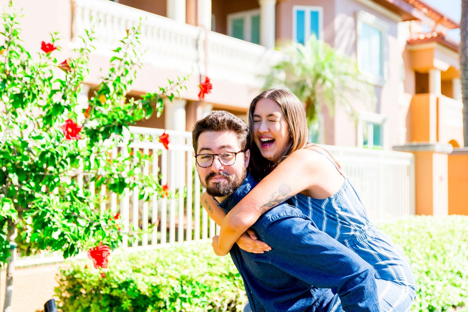 young woman piggyback on boyfriend, laughing next to pink hibiscus tree at Vinoy Park sunrise engagement pictures