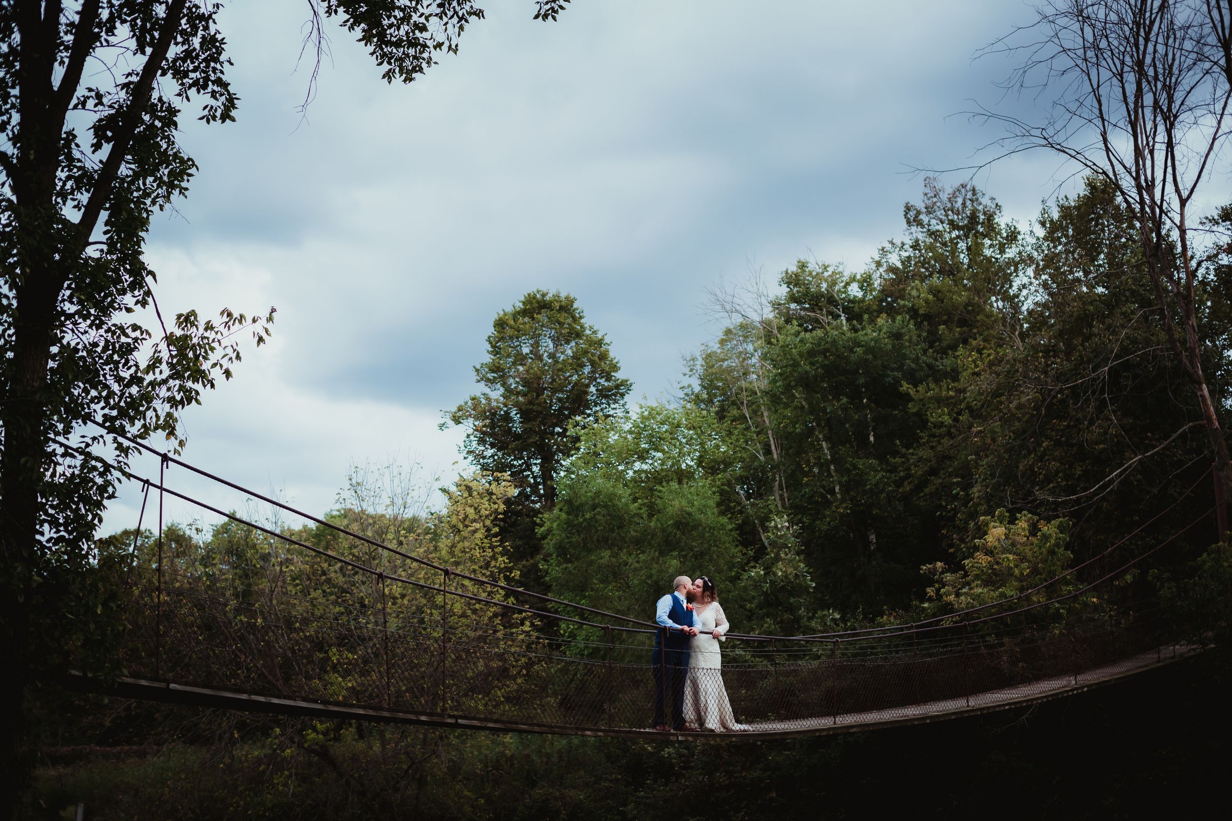 Man and woman in wedding attire kissing in the middle of a swinging cable rope bridge.