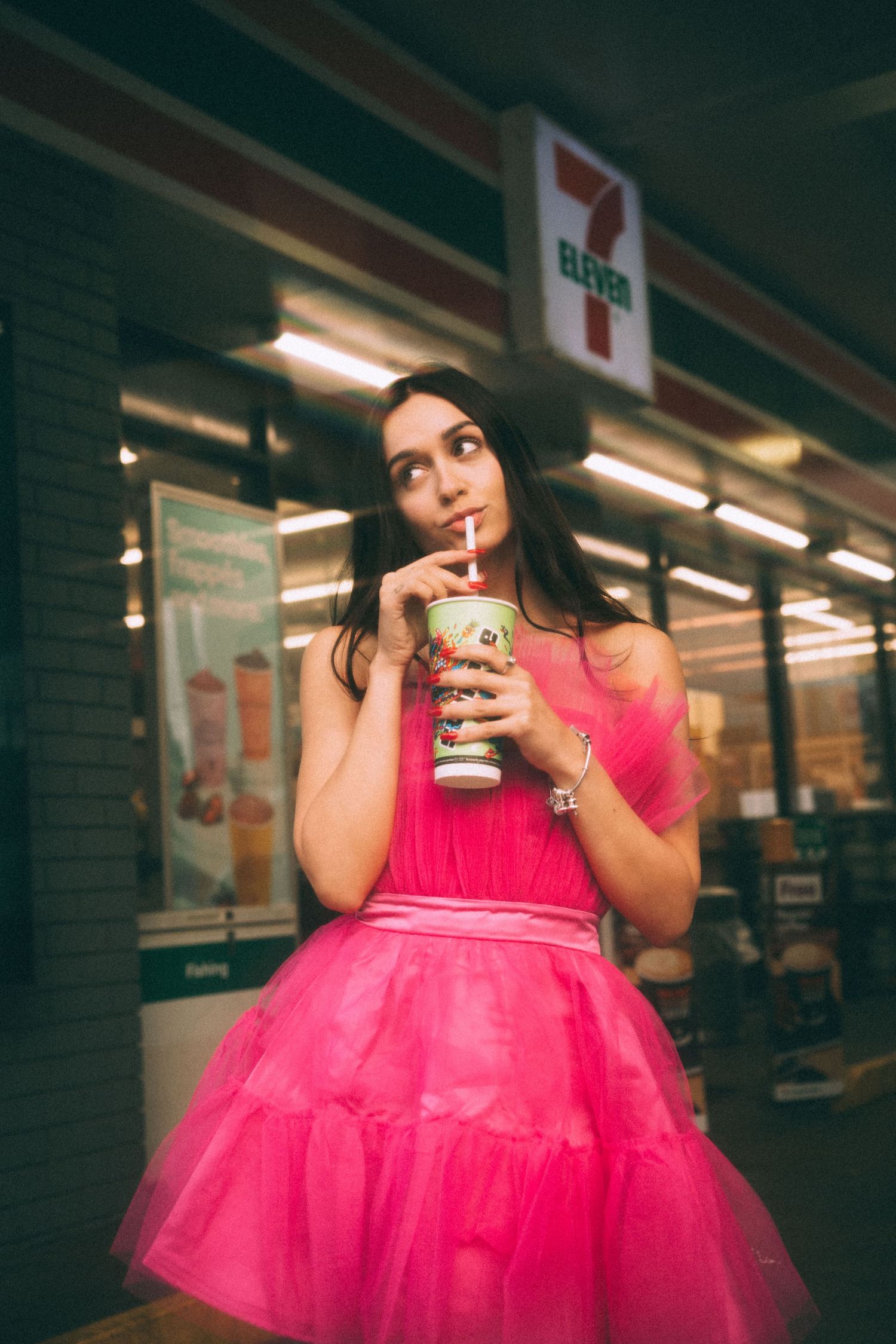 Someone in a vibrant pink tulle dress sips a drink outside a 7-Eleven store at night.